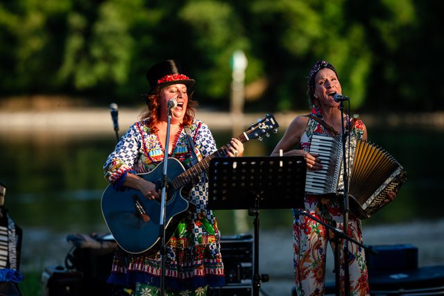 Die zwei Schrullen bei der Beachparty «Helft uns helfen» in Hemishofen 2026: eine mit Zylinder und Gitarre, die andere mit Akkordeon, beide singen, dahinter Wasser und Bäume